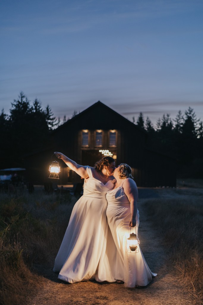 Jessica Clark and her wife kissing outside a lit barn at dusk on their wedding day, each holding a lantern